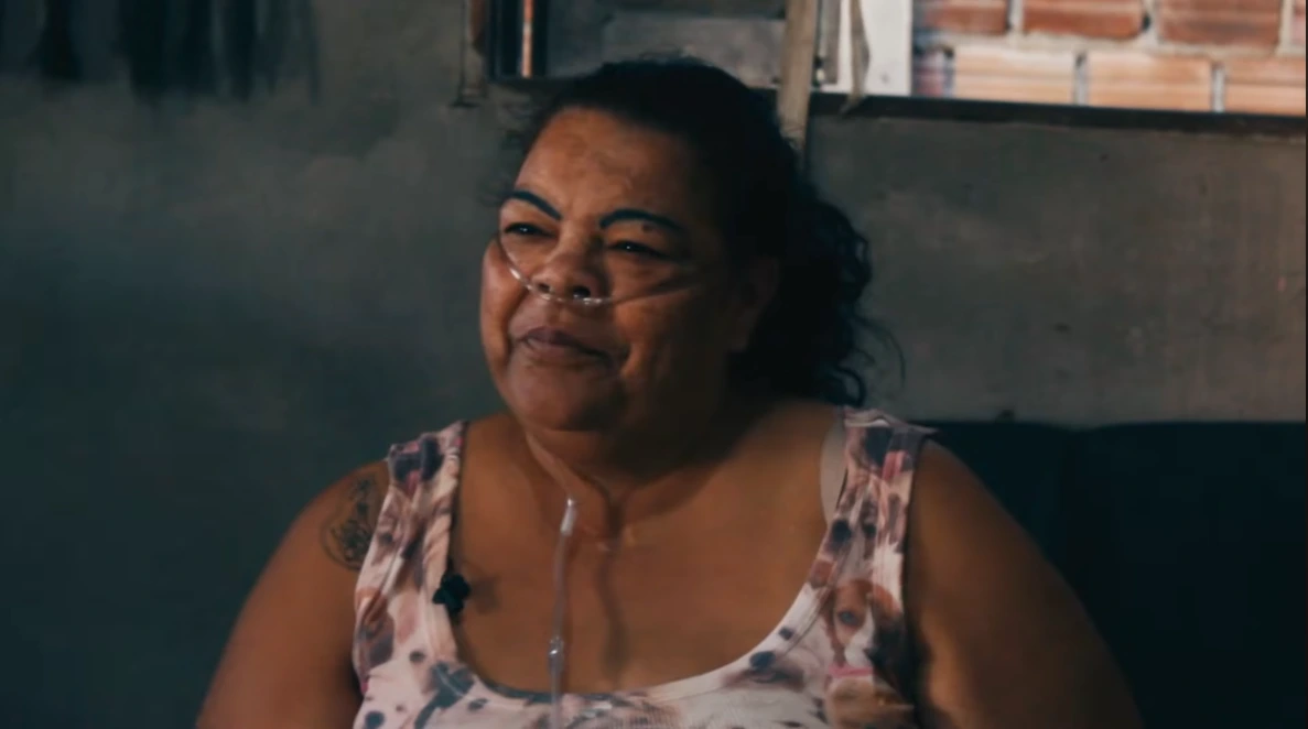 Foto de Maria Gorete sorrindo em sua casa, vestida com uma blusa com rosas e tubo respiratório no nariz.